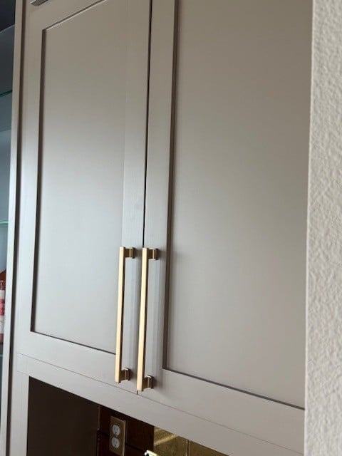 Two white cabinet doors with brass cylindrical handles mounted on a beige wall above a dark lower cabinet with electrical outlet